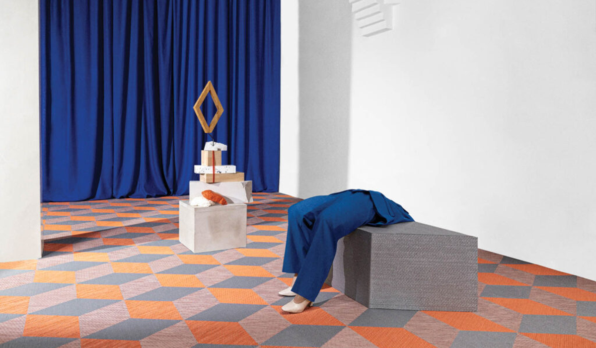 person hunched over grey block on kaleidoscopic flooring against blue drapes