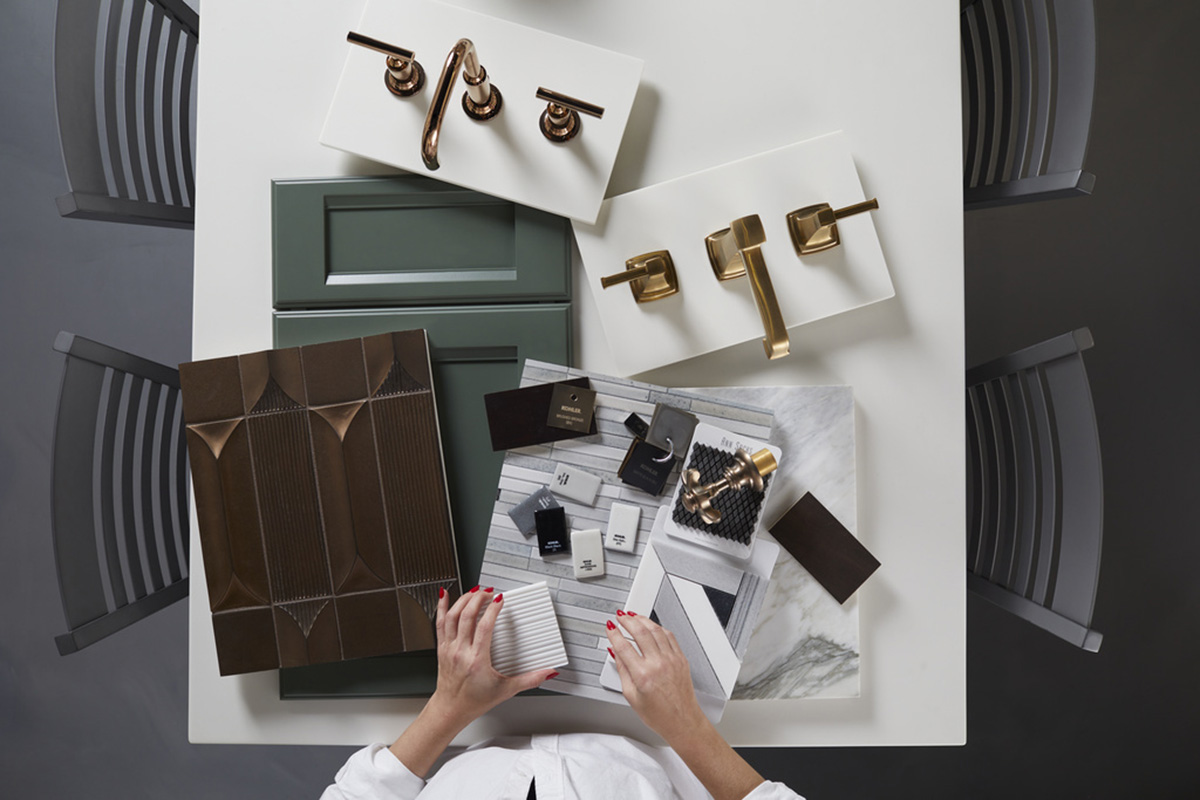 An assortment of samples — tiles, faucets, panels — are displayed on a table while a woman with a red manicure picks one up.